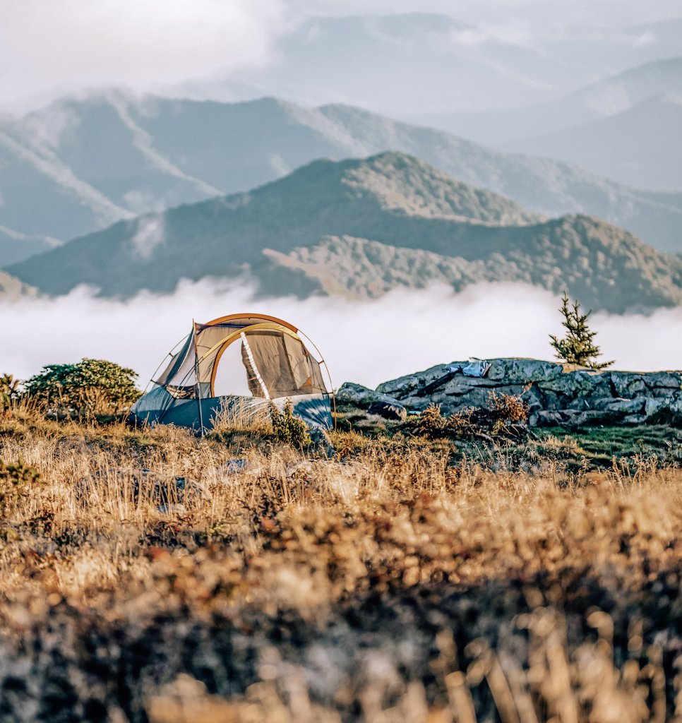 A tan colored tent alone in a field in front of mountains.