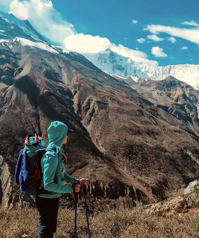 Girl trekking in Nepal's mountains in a blue jacket, backpack, and trekking poles - three of the Nepal trekking essentials on your packing list.