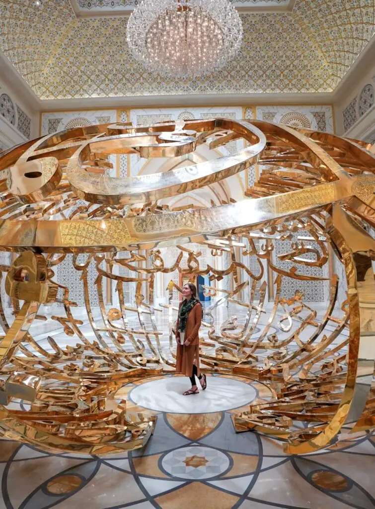 Monica underneath a gold sculpture at Qasr Al Watan, a beautiful place to explore when Visiting Abu Dhabi as a Woman.