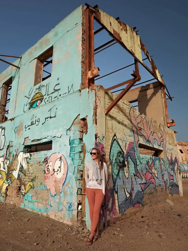 Girl in front of an abandoned building in Kuwait, wearing long pants, a flowy white top and scarf.