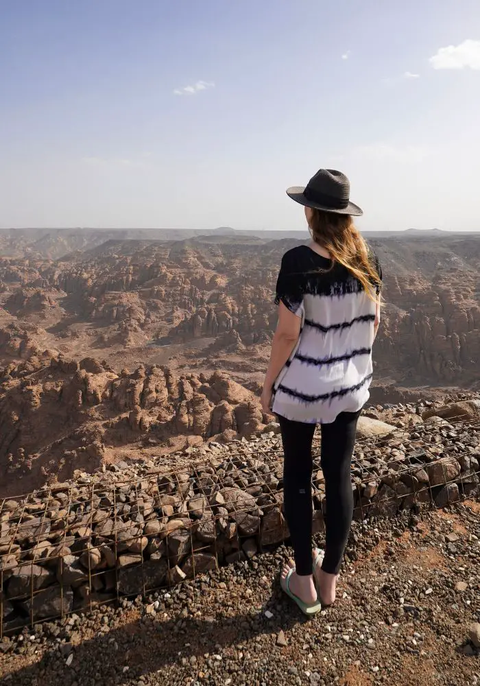 Monica looking out at AlUla from the Herrat Viewpoint, wearing a black hat and black and white top.