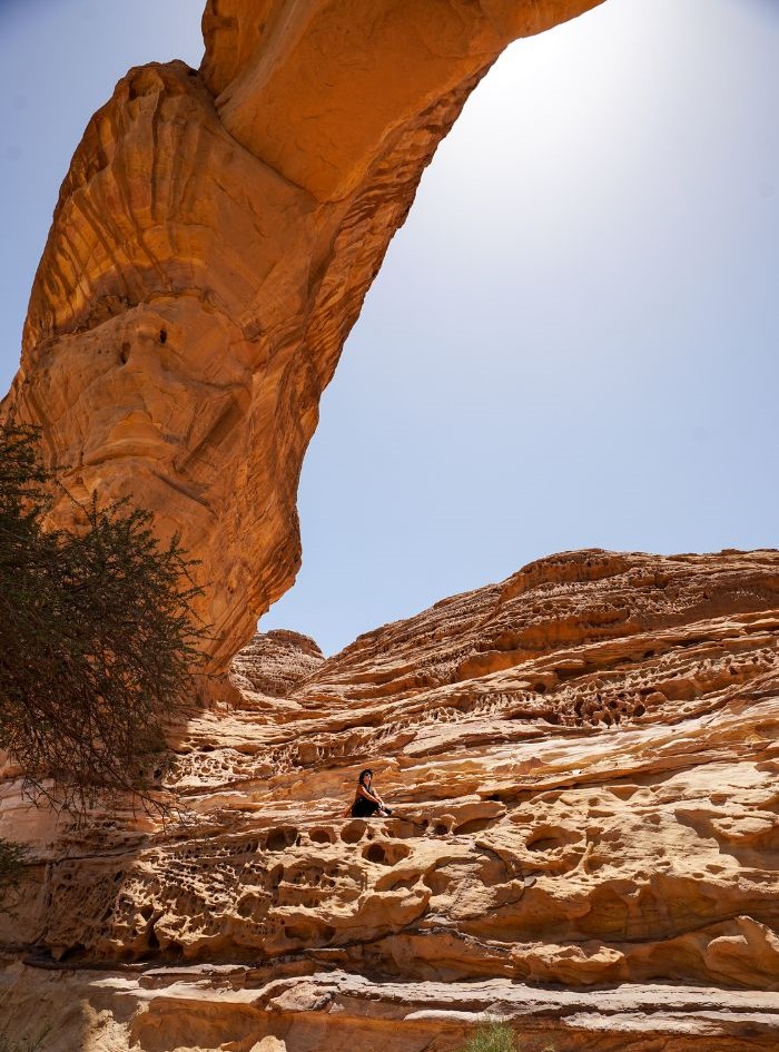 Monica underneath an orange rock archway in Saudi Arabia, where she travels as an American, from a distance.