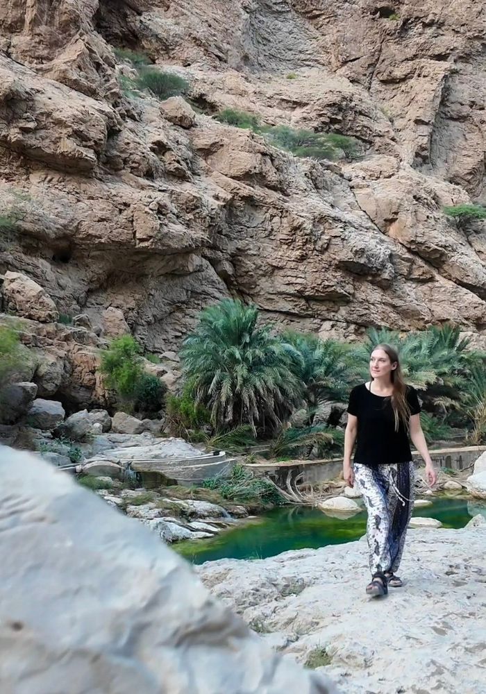 Monica wearing a black shirt and loose white pants, walking through the mountainous Wadi Shab - one of the best day trips from Muscat, Oman.