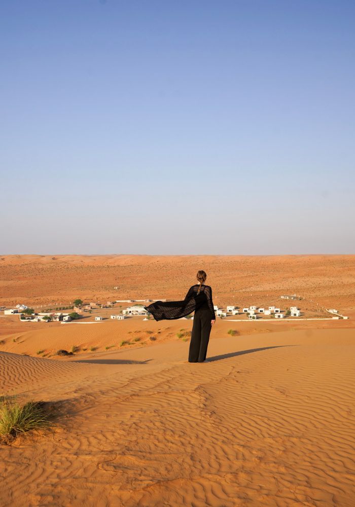 Monica standing on the orange Oman sand, looking out to a white desert camp.