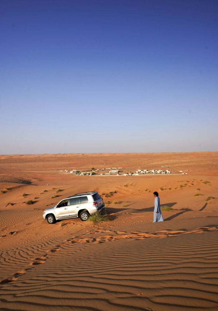 The patterned orange sand dunes in Wahiba Sands on a bright sunny day, with a 4X4 car and a small camp in the distance. 