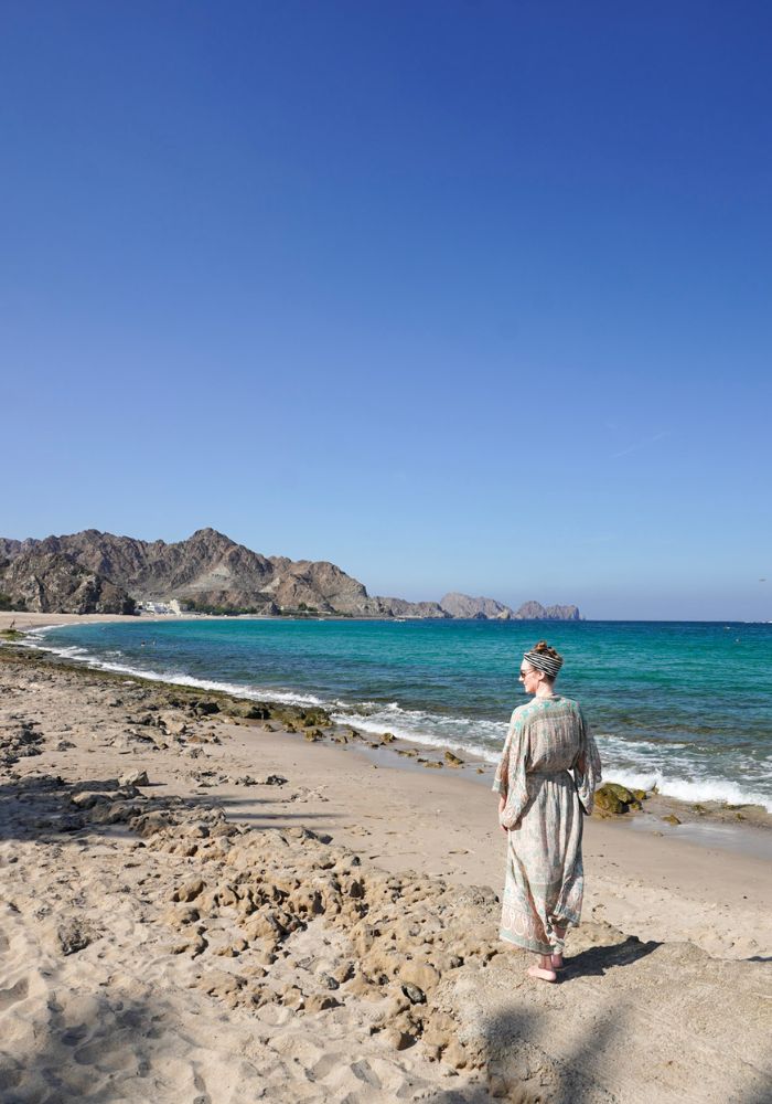 Monica standing on the shore of Al Bustan Beach, one of Muscat's best beaches, with blue water and rugged mountains.